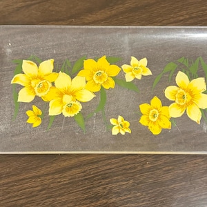 May include: Rectangular clear glass tray with a floral design. The tray features yellow daffodils with green leaves. The flowers are arranged across the tray's surface. The tray is set on a dark brown wooden surface.
