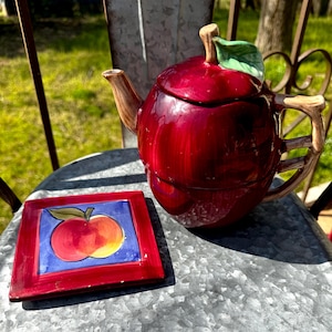 May include: A red apple-shaped teapot with a green leaf and brown stem, and a matching square plate with an apple design. The teapot and plate are on a metal surface, with a blurred green background.