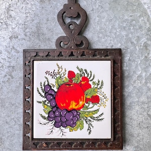 Puede incluir: Un salvamanteles vintage con un diseño de frutas. La baldosa de cerámica cuadrada presenta una manzana roja, uvas moradas y fresas rojas rodeadas de hojas verdes. La baldosa está enmarcada en un marco de metal marrón oscuro con un asa ornamentada.