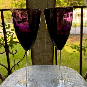 May include: Two wine glasses featuring deep purple, translucent bowls and clear, slender stems with circular bases. The glass bowls have a smooth, tapered shape, and the clear stems provide a contrast to the dark, rich color of the upper portion.