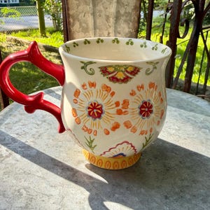 May include: A ceramic mug with a cream-colored base featuring orange and red floral patterns. The mug has a bright red, uniquely shaped handle and a yellow base. The interior rim is decorated with a repeating pattern of small green dots.