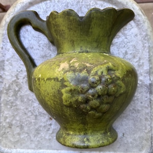 May include: A moss green ceramic pitcher featuring a scalloped rim, a curved handle, and a raised relief design of a grape cluster with leaves on the front. The surface has a distressed, weathered finish with visible patches of lighter tan color.