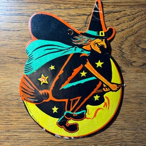 May include: Vintage Halloween decoration of a witch on a broomstick against a yellow crescent moon. The witch is in black with orange and green details, and the moon has yellow stars. The decoration is made of cardboard.