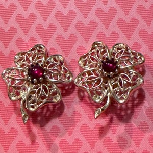 May include: A pair of gold-toned clover-shaped earrings with a filigree design. Each earring features a round, deep red gemstone at the center. The earrings are set against a pink background with a heart pattern.