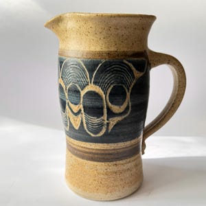 May include: A stoneware pitcher with a tan and dark blue design. The pitcher has a spout and a handle. The dark blue section features a repeating pattern of stylized, interconnected shapes. The top and bottom sections are tan with a speckled texture.