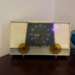 May include: Vintage Philco clock radio with a cream-colored plastic casing and a dark blue clock face. The clock features a starburst design and yellow knobs for volume and tuning. The clock measures approximately 10 inches wide.
