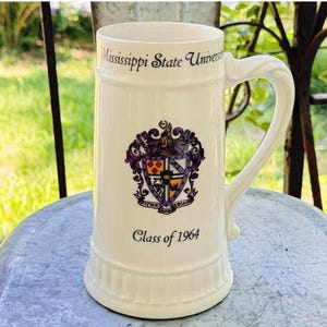 May include: Cream-colored ceramic beer stein with a large handle. The stein features the words "Mississippi State University" and "Class of 1964". A colorful emblem is centered on the stein.
