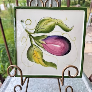 May include: Square ceramic tile with a hand-painted eggplant design. The eggplant is purple and pink, with green leaves and a green border. The tile is displayed on a decorative metal stand.