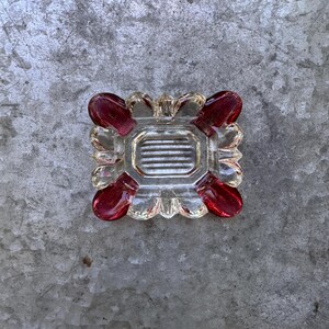 May include: A clear glass ashtray with a square shape and a central rectangular area with horizontal lines. The corners feature red, rounded accents. The ashtray is designed for holding cigarettes or cigars.