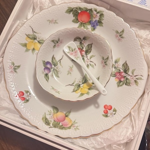 Peut inclure: Un ensemble de vaisselle en porcelaine blanche avec un motif de fruits, comprenant une grande assiette, une assiette plus petite, un bol et une cuillère. L'ensemble présente des illustrations de pêches, de citrons, de prunes et de cerises avec des feuilles vertes et des fleurs roses. Les bords sont dorés.