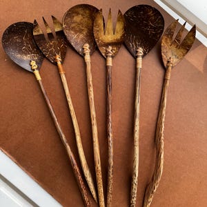 May include: Set of six handcrafted salad servers. The serving spoons and forks are made from coconut shells and wood. The spoons have a rounded bowl shape, while the forks have three tines. The handles are made of wood and are connected to the serving heads with woven twine.