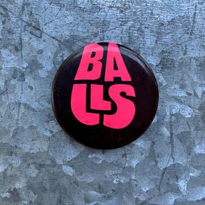 May include: A round, black button with the word "BALLS" in bright pink, bold letters. The button is centered against a textured, metallic gray background.