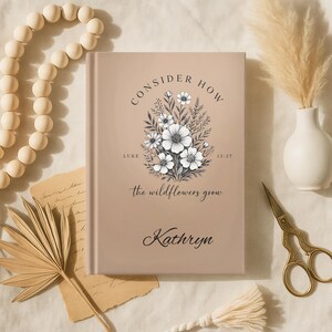 May include: A beige journal with the text "CONSIDER HOW" and "the wildflowers grow." A floral illustration with white flowers and green leaves is centered on the cover. The name "Kathryn" is written below the text. Decorative items surround the journal.