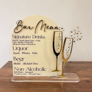 May include: A white acrylic sign with black text listing a bar menu. The sign features a gold silhouette of two champagne glasses with gold bubbles. The menu includes signature drinks, liquor, beer, and non-alcoholic options.