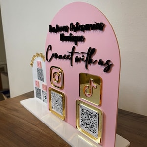 May include: Pink and white sign with the text "Accessories Boutique, Connect with us" in black lettering. The sign features icons for Instagram, TikTok, and QR codes.