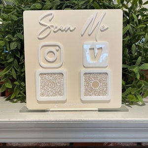 May include: A white sign with the text "Scan Me" in white lettering. The sign has two QR codes, one for Instagram and one for TikTok, with the logos for each platform above the codes.