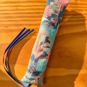 Può includere: Un giocattolo per gatti colorato e fantasia con nastri blu. Il tessuto presenta un mix di disegni astratti rosa, verde acqua e blu navy. L'etichetta recita "SECOND WIND SENDS". Il giocattolo è su una superficie di legno.