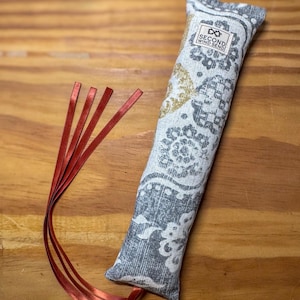 May include: A rectangular fabric toy featuring a gray and white paisley pattern with gold accents. A white label with the text SECOND WIND SEND is stitched near the top. Four thin, metallic red ribbons are attached to the bottom corner.