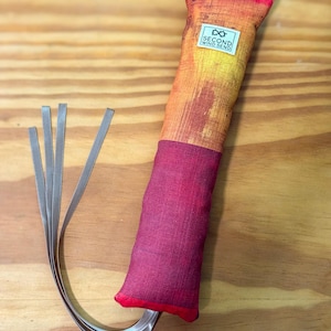 May include: A rectangular fabric toy featuring a gradient of orange and yellow on the top half and a solid deep red on the bottom half. A white rectangular label with the text SECOND WIND SEND is attached. Four thin grey ribbons extend from the base.