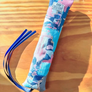 Könnte beinhalten: Ein langes, rechteckiges Katzenspielzeug mit einem farbenfrohen, abstrakten Muster in Blau-, Rosa- und Weißtönen. Das Spielzeug hat ein kleines Etikett mit der Aufschrift "SECOND WIND SEND" und ist mit drei blauen Bändern versehen.