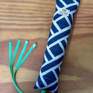 May include: A rectangular fabric toy featuring a dark blue and white geometric lattice pattern. A small white label with the text SECOND WIND SEND is stitched near the top. Four thin, bright neon green ribbons extend from the bottom end.