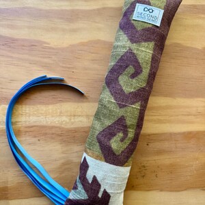 May include: A long, rectangular cat toy with a patterned fabric in olive green and burgundy. The fabric features a geometric design. The toy has a white tag with the text "SECOND WIND SEND". A blue ribbon is attached to the end.