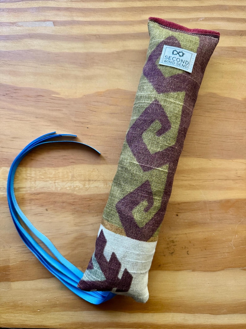 May include: A long, rectangular cat toy with a patterned fabric in olive green and burgundy. The fabric features a geometric design. The toy has a white tag with the text "SECOND WIND SEND". A blue ribbon is attached to the end.