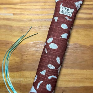 May include: A rectangular fabric pouch in a deep reddish-brown color with a white abstract leaf pattern. A white rectangular label with the text SECOND WIND SEND is stitched near the top. Three thin, light blue ribbons extend from the bottom.