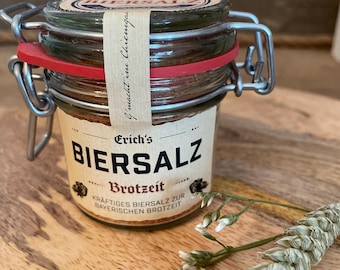 May include: A small glass jar of "Biersalz" with a metal clasp and a red rubber seal. The label features the text "Erich's Biersalz Brotzeit" and "Kräftiges Biersalz zur bayerischen Brotzeit." A sprig of wheat and small white flowers are in the foreground.