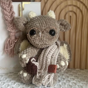 May include: A plush crochet dragon toy made of soft taupe yarn with cream-colored accents on the horns, wings, and feet. The dragon wears round gold-rimmed glasses and a long, fringed cream-colored scarf, and holds a small dark brown crocheted book.