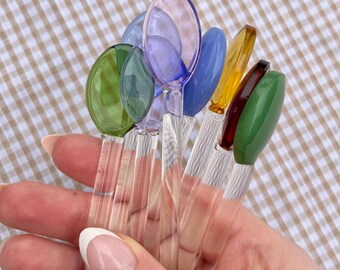 May include: A set of six glass spoons with clear handles and colorful, rounded bowls. The bowls are in shades of green, blue, purple, and yellow.