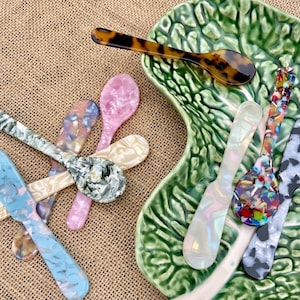 May include: A selection of decorative spoons in a variety of colours and patterns, including pink, blue, and tortoiseshell. The spoons are displayed on a green, leaf-shaped plate and a burlap surface.