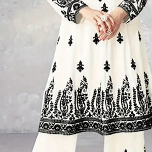 Könnte beinhalten: Weißes zweiteiliges Outfit mit aufwendiger schwarzer Stickerei. Das langärmlige Oberteil und die weite Hose weisen detaillierte Blumen- und Paisley-Muster am Ausschnitt, an den Ärmeln und am Saum auf. Traditioneller Stil.