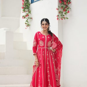 Könnte beinhalten: Eine Frau trägt ein rosa-goldenes besticktes Kleid mit passendem Dupatta. Das Kleid hat einen ausgestellten Rock und ein tailliertes Oberteil. Die Frau steht vor einer weißen Wand mit einem blauen Geländer.