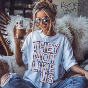 May include: A white t-shirt with the text "THEY NOT LIKE US" in silver glitter letters with a purple outline. The shirt is being worn by a person with blonde hair and sunglasses.