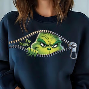 Könnte beinhalten: Ein dunkelblaues Sweatshirt mit einer Grafik eines grünen, pelzigen Wesens, das durch einen offenen Reißverschluss schaut. Das Wesen hat einen mürrischen Ausdruck und eine Hand ist sichtbar. Der Text "EW PEOPLE" steht in grünen Buchstaben daneben.