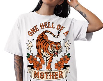 May include: White t-shirt with a graphic design featuring a tiger, orange flowers, and the text "ONE HELL OF A MOTHER" above "FERAL MOMS CLUB". The tiger is orange and black, with a fierce expression.