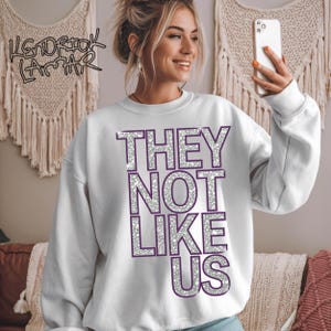 May include: White sweatshirt with the text "THEY NOT LIKE US" in silver glitter letters with a purple outline.