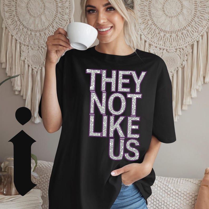 May include: Black t-shirt with the text "THEY NOT LIKE US" in silver glitter letters with purple outlines.