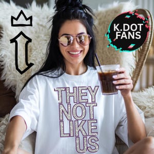 May include: White t-shirt with the text "THEY NOT LIKE US" in silver glitter letters. The letters have a purple outline.