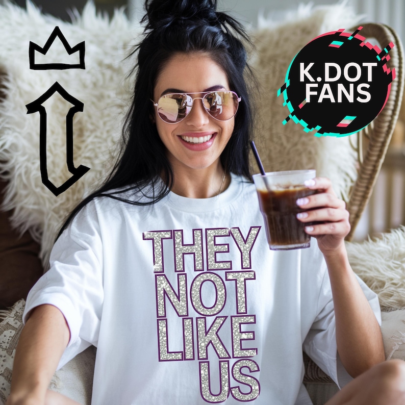 May include: White t-shirt with the text "THEY NOT LIKE US" in silver glitter letters. The letters have a purple outline.