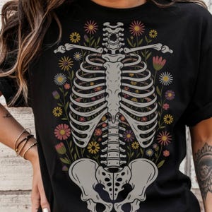 May include: Black t-shirt with a white skeleton design. The skeleton is surrounded by colorful flowers.