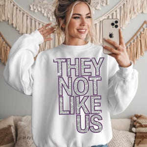 May include: White sweatshirt with the text "THEY NOT LIKE US" in silver glitter letters with a purple outline.