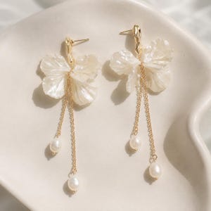 May include: A pair of gold-tone drop earrings featuring translucent, off-white, petal-shaped components attached to textured gold posts. Each earring includes two delicate gold chains of varying lengths, each finished with a small white pearl.