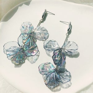 Puede incluir: Un par de pendientes colgantes de flores de color azul iridiscente con ganchos plateados. Los pendientes están hechos de plástico transparente y tienen un aspecto delicado y femenino.