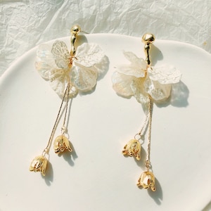 Puede incluir: Un par de pendientes de tono dorado con abalorios en forma de flor blanca y campanillas doradas colgantes. Los pendientes están sobre una superficie blanca.