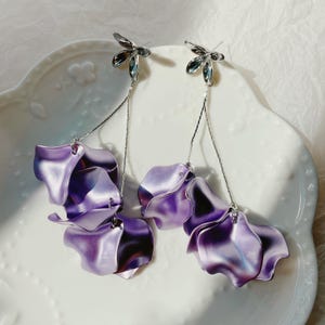 May include: A pair of dangling earrings featuring a floral design. Each earring has a silver-toned stud with a three-petal design, connected to a chain that suspends three large, iridescent purple petals. The earrings are displayed on a white, decorative plate.
