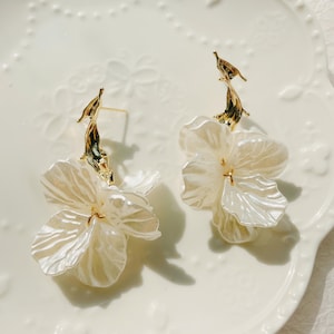 May include: A pair of earrings with large, pearlescent white flower pendants. The earrings have gold-coloured, abstract post attachments. The flowers have multiple petals and a subtle sheen, creating a delicate and feminine aesthetic.