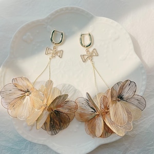 May include: A pair of gold-toned drop earrings. Each earring features a small bow-shaped accent at the top, connected to a cluster of layered, wire-like flower petals. The earrings are displayed on a white, decorative plate.