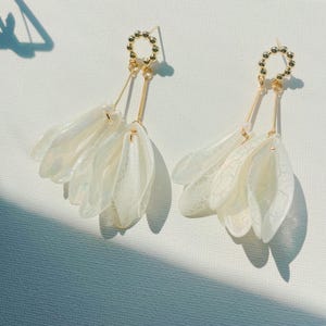 Puede incluir: Un par de pendientes de oro con abalorios en forma de flor blanca e iridiscente. Los pendientes tienen un diseño delicado y femenino.